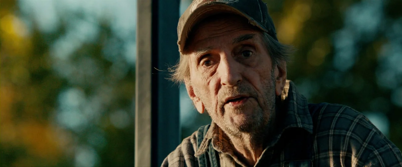 The Last Stand (2013) movie still: closeup — An elderly man in a baseball cap stands outdoors during the late afternoon. He is talking to someone…