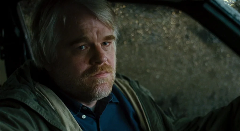 The Savages (2007) movie still: closeup — The interior of a car features dark upholstery and an olive green hooded jacket. A middle-aged man with…