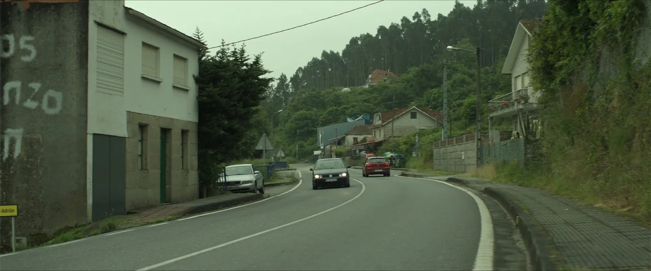 Apocalypse Z: The Beginning of the End (2024) movie still: wide — A rural asphalt road winds between a hillside lined with houses and vegetation.…
