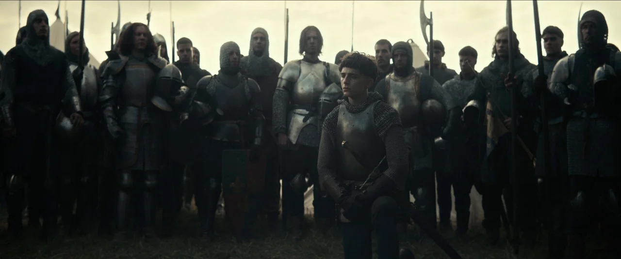 The King movie still: fullbody — A field filled with rows of soldiers in medieval plate armor and chainmail hoods. A young knight kneels on the…