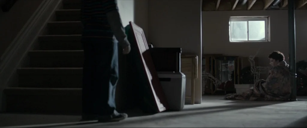 Clown (2014) movie still: wide — A dimly lit basement features concrete floors, exposed ceiling joists, and a small, high window. A young child…