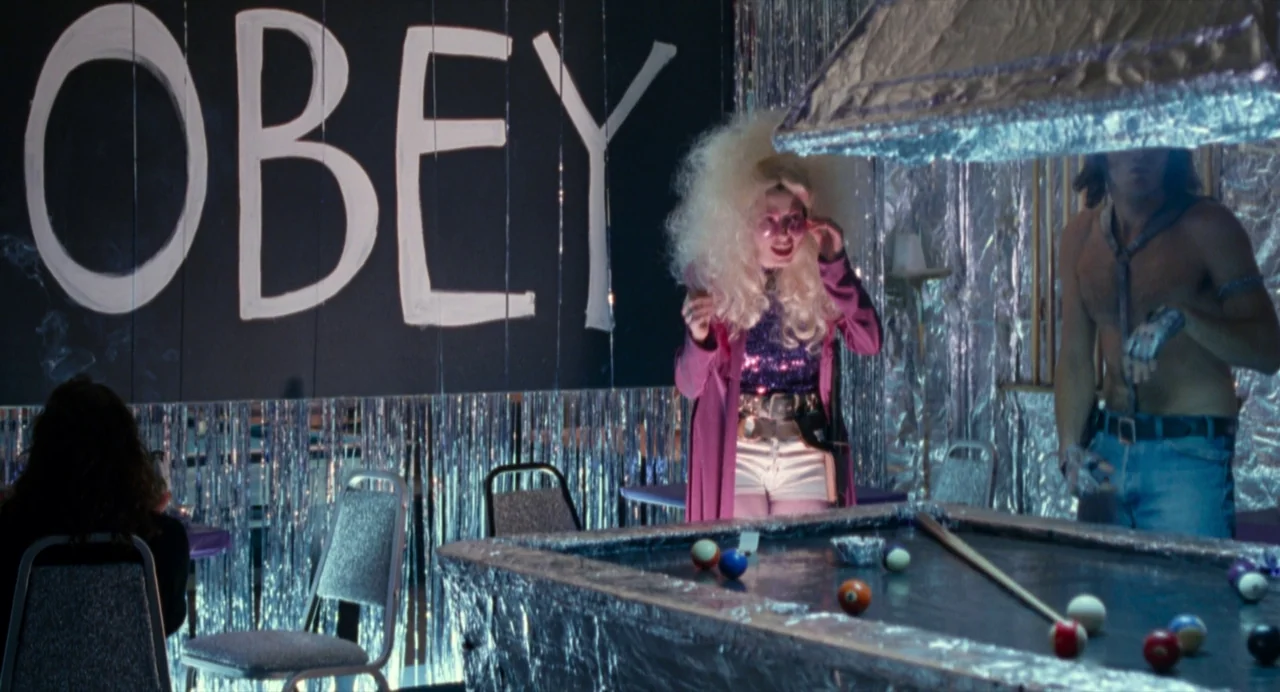 The Doom Generation (1995) movie still: wide — A basement room is decorated with silver tinsel curtains and a large black Obey sign. A woman with a…