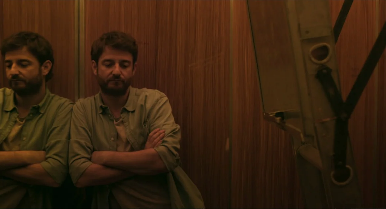 The Lift (2021) movie still: medium — A confined elevator interior features dark wood-paneled walls and a metallic corner fixture.