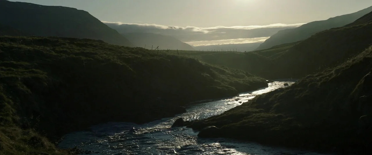 Babel (2006) movie still: establishing — A wide river winds through a deep, grassy mountain valley under a bright horizon. The sun silhouettes the…