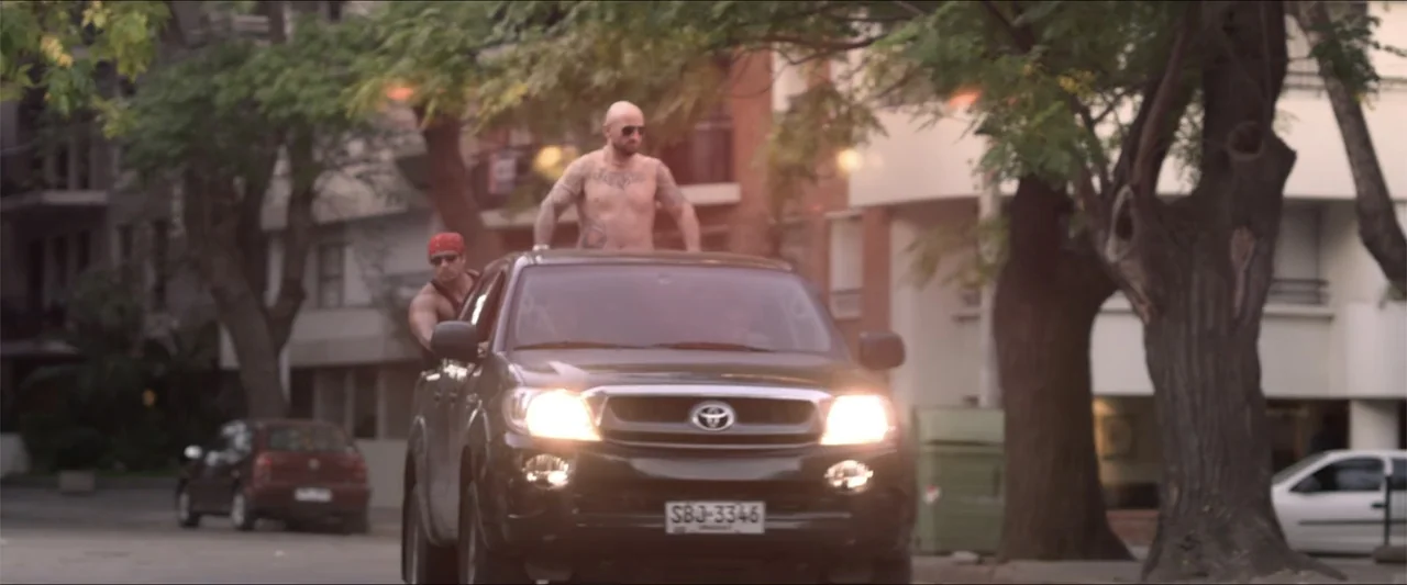 High Five (2004) movie still: medium — A dark pickup truck moves forward on a paved road flanked by tall trees. Two men ride the vehicle with one…