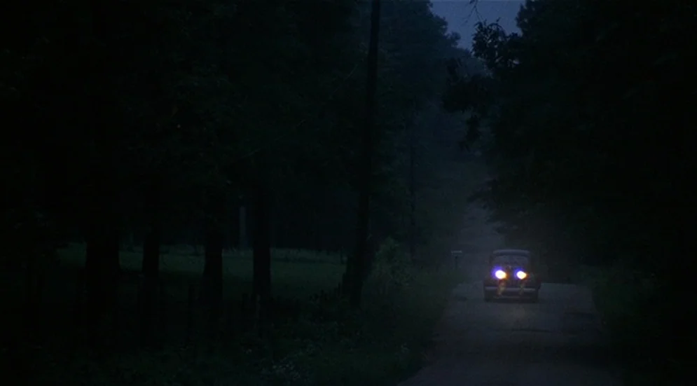 Thieves Like Us (1974) movie still: wide — A narrow road is flanked by dark dense woods under a low-light night sky. A dark sedan approaches from…