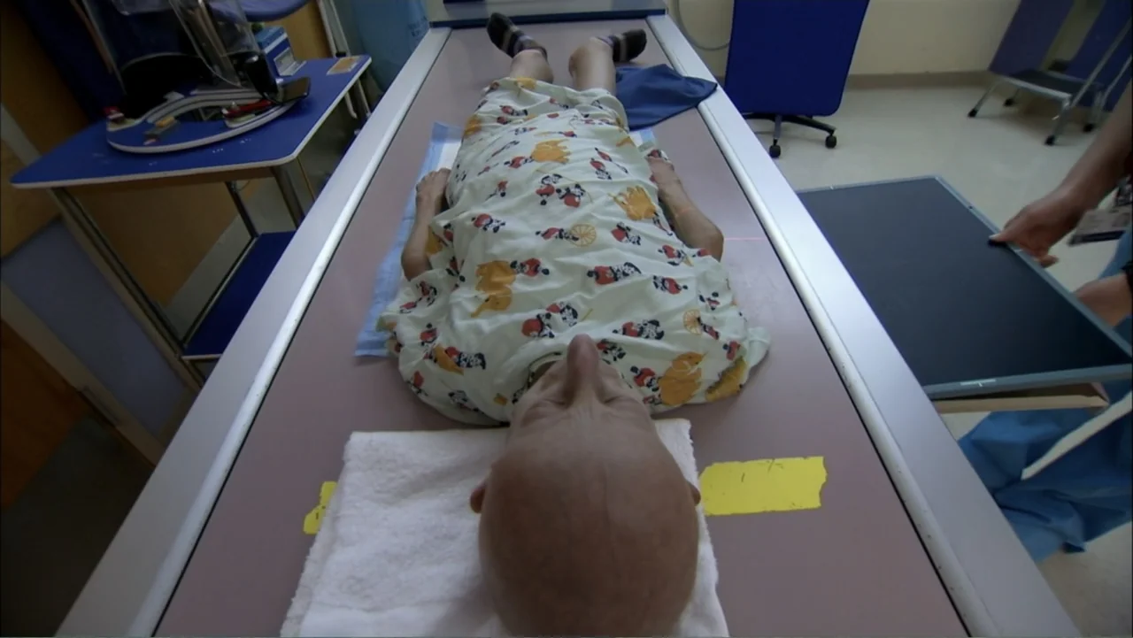 Life According to Sam (2013) movie still: medium — A clinical examination room features a metallic table under bright overhead fluorescent lights. A…