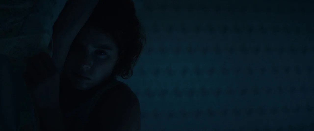 Beast (2017) movie still: closeup — A dark bedroom with minimal light reflecting on a surface. A person lies in bed with only their face partially…