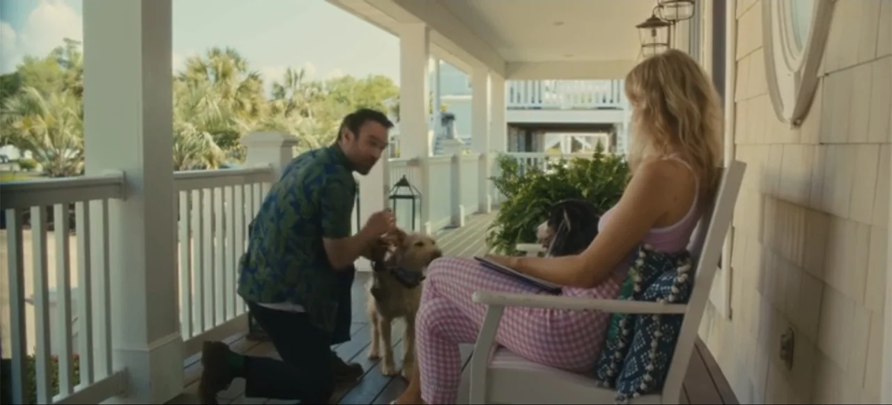Merv (2025) movie still: medium — A sunny residential porch with white railings and wood flooring serves as the setting. A man kneels to pet a dog…
