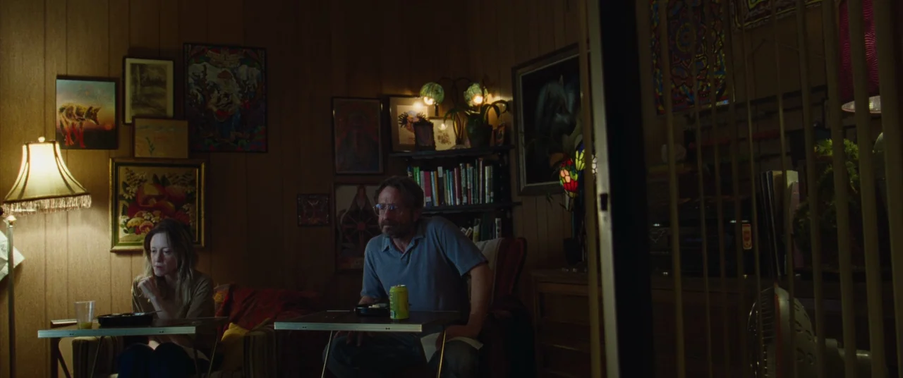 To Leslie (2022) movie still: medium — A dimly lit room with wood-paneled walls decorated with multiple framed paintings and prints. A man and woman…