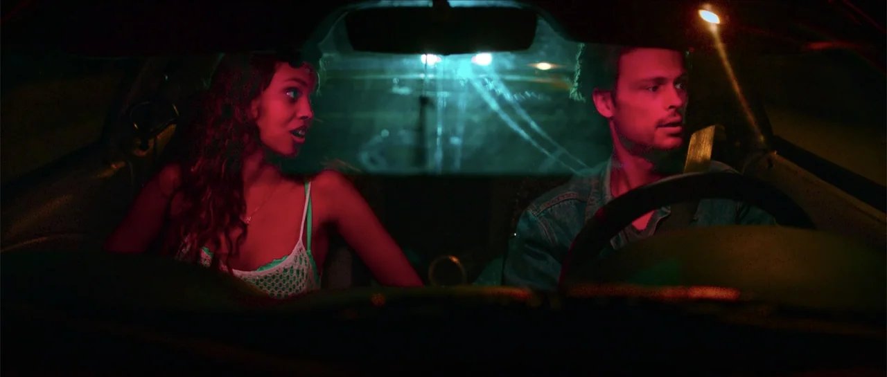 68 Kill (2017) movie still: medium — The interior of a moving car at night is illuminated by harsh, colorful light. A man drives while a woman looks…