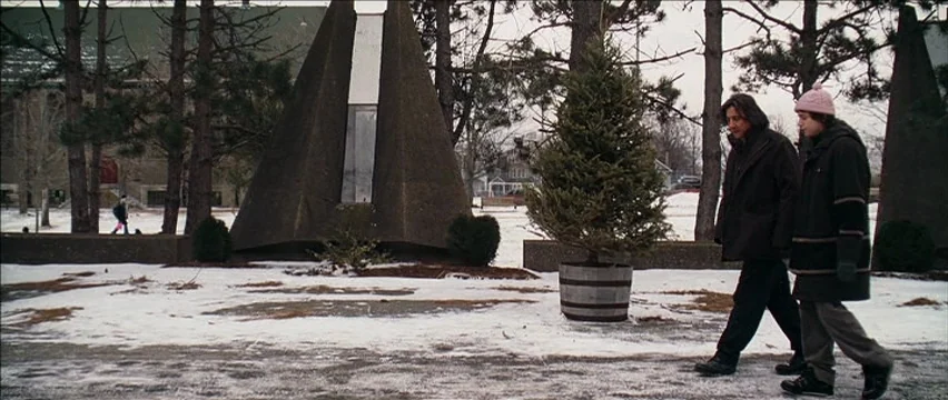 Snow Angels (2007) movie still: fullbody — A snow-covered public space features modern triangular concrete architecture and a potted pine tree. Two…