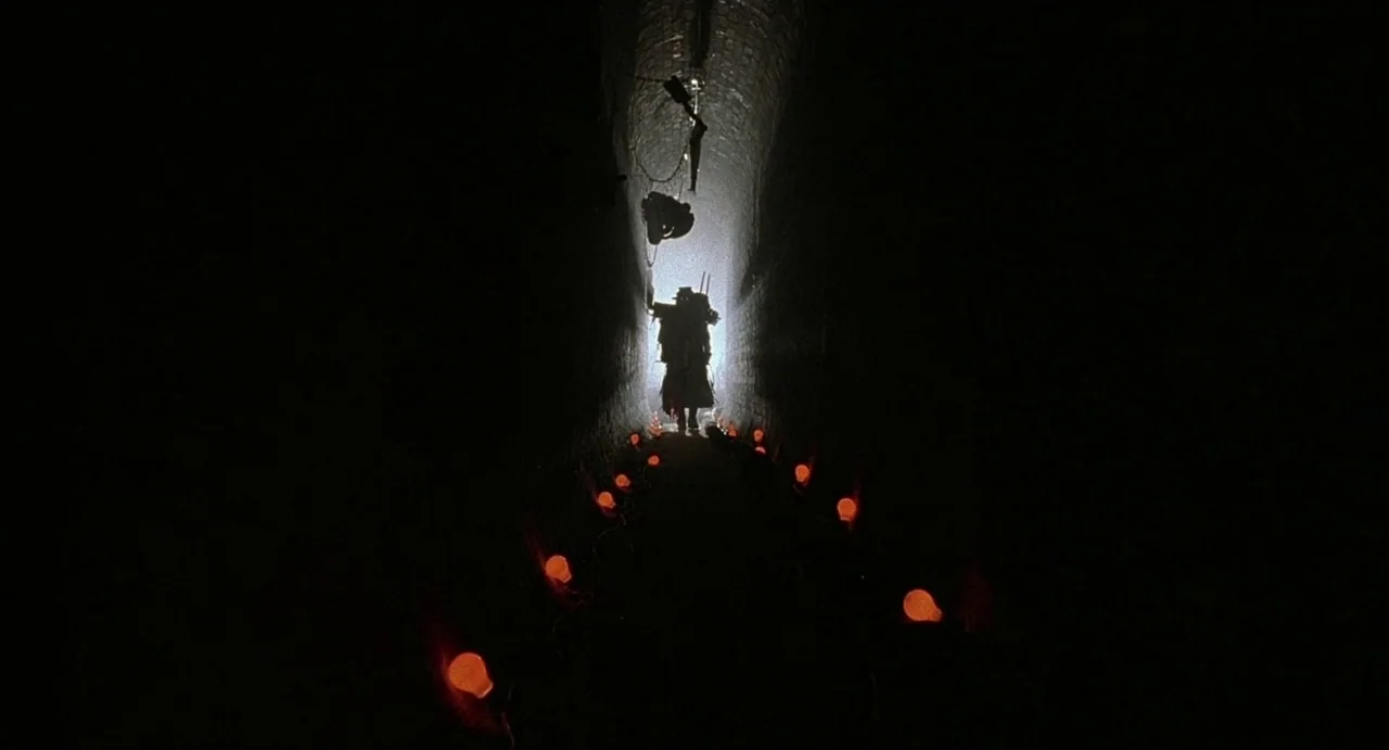 Hardware (1990) movie still: wide — A long dark brick tunnel is lit by a line of glowing orange bulbs. A single silhouetted figure in an overcoat…