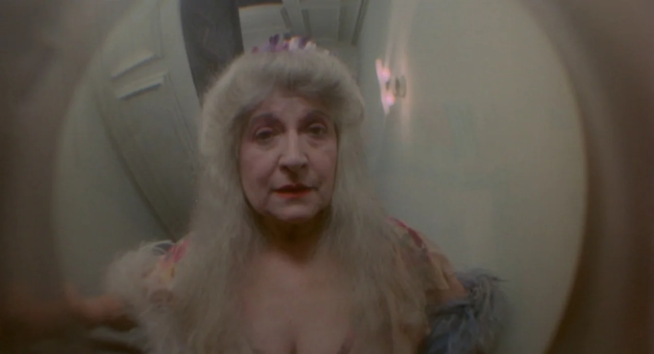 Ms. 45 (1981) movie still: closeup — An elderly woman stands before a wide-angle door peephole in a hallway. The circular frame distorts the view of…