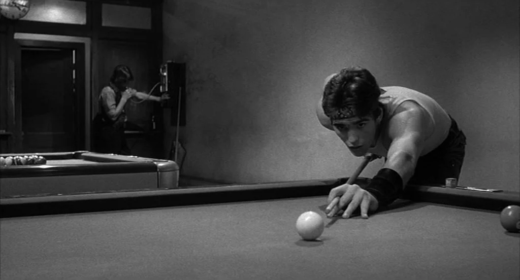 Rumble Fish (1983) movie still: medium — A dimly lit room features a dark table surface and a wall-mounted telephone. A man in a tank top prepares…