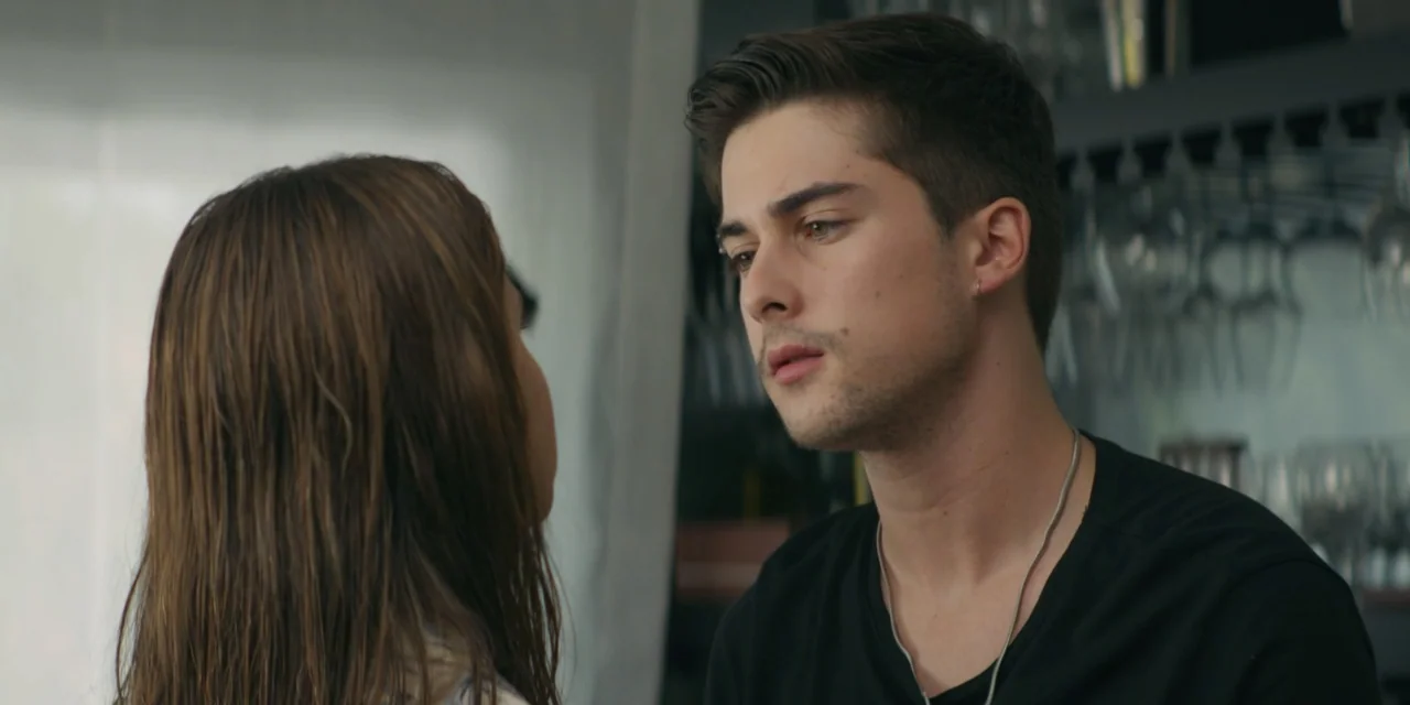 My Fault (2023) movie still: medium — A man and woman face each other in an interior room with visible glassware racks. The man watches the woman…