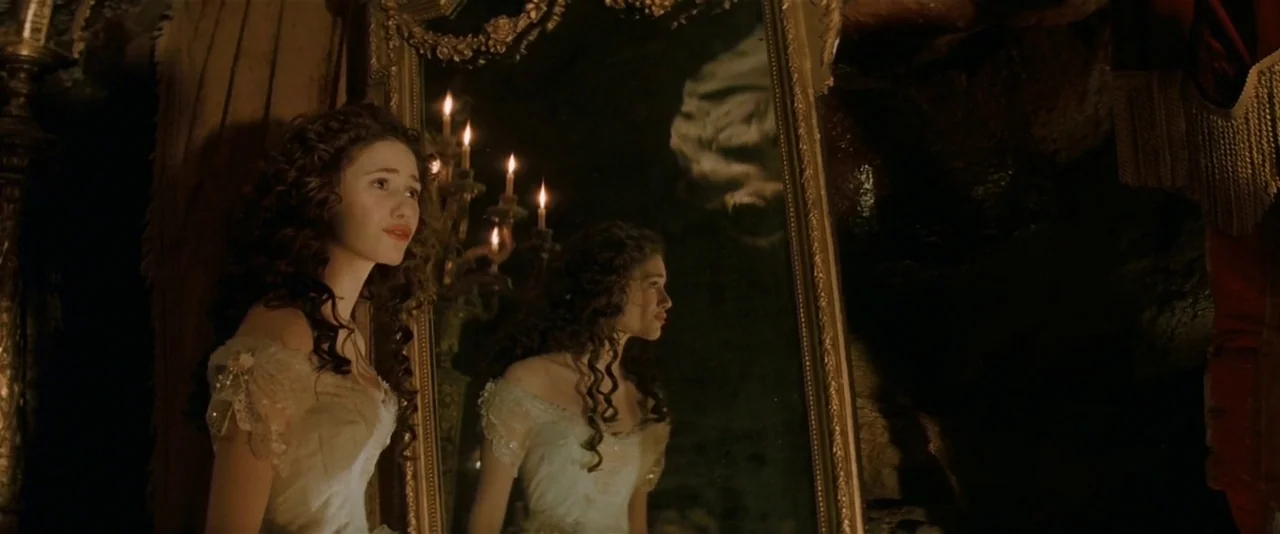 The Phantom of the Opera (2004) movie still: medium — An ornate gold-framed mirror stands against a dark, rough-hewn stone wall in a room. A young…