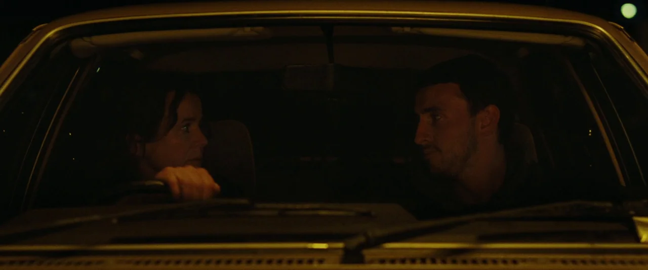 God’s Creatures (2022) movie still: medium — The interior of a car at night is mostly obscured by deep shadows. A woman drives while a man sits in…