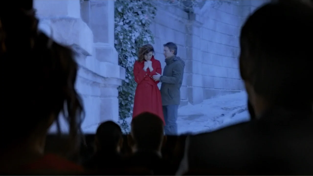 I Married a Dumbass (2016) movie still: wide — Audience members sit in a dark room watching a screen projecting a film. A woman in a red coat and a…