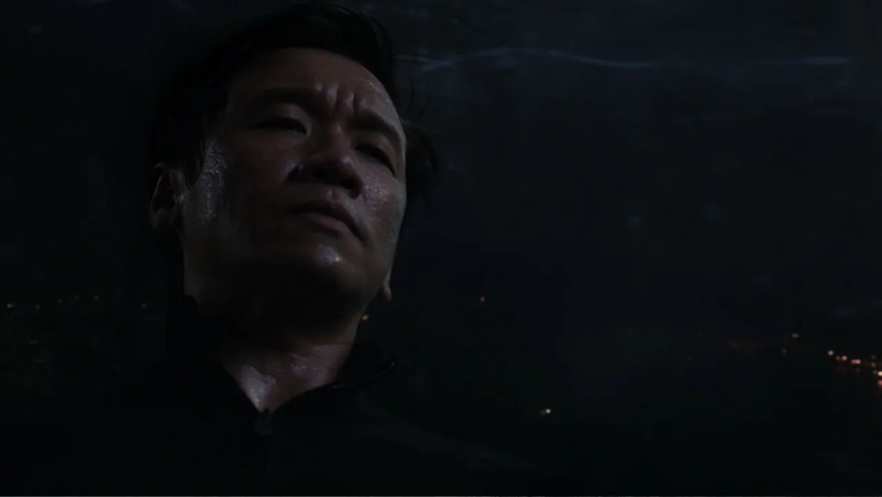 Skyscraper (2018) movie still: closeup — A man stands in a dark outdoor environment at night with distant city lights. The man gazes upwards with a…