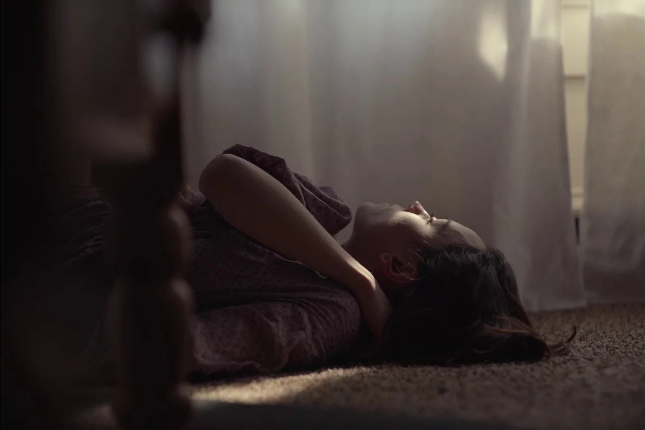 Sometimes I Think About Dying (2023) movie still: medium — A carpeted floor lies beneath a person in a room with white sheer curtains. A person…