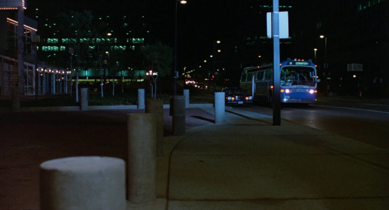 Polyester (1981) movie still: wide — An empty urban sidewalk features a row of cylindrical concrete bollards at night. A municipal bus drives down…