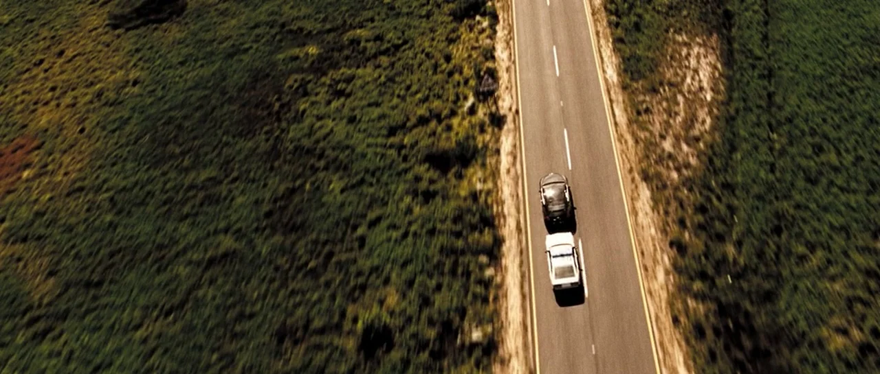 Doomsday (2008) movie still: wide — An asphalt road cuts through a field covered in green scrub vegetation. Two vehicles drive down the straight…