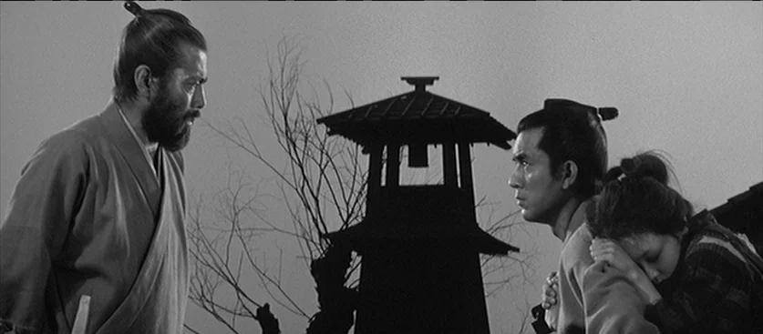 Red Beard (1965) movie still: medium — An open village area features a central wooden fire-watch tower and leafless trees. Two men face each other…
