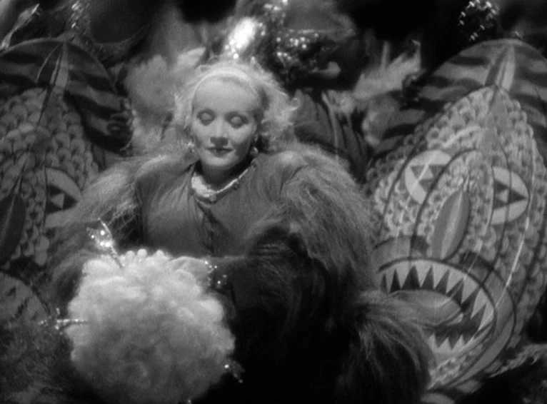 Blonde Venus (1932) movie still: medium — A theatrical stage set features large decorated oval shields flanking a central female figure. The woman…