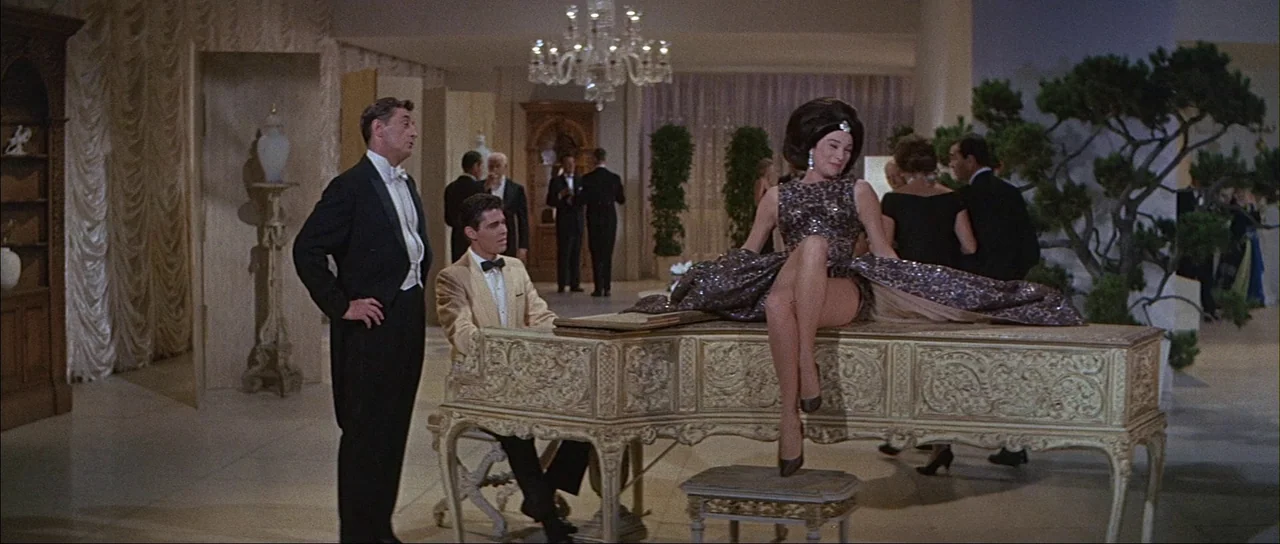 What A Way To Go! (1964) movie still: fullbody — A spacious ballroom with ornate beige decor, crystal chandeliers, and a white grand piano. A woman…
