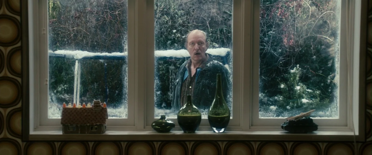 The Box (2009) movie still: medium — An older man stands outside a house looking through a large glass window. A gingerbread house and several glass…