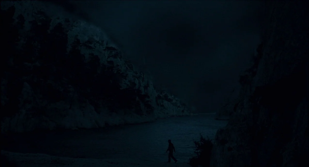 The Army of Shadows (1969) movie still: wide — A dark rocky cove is flanked by steep cliffs under a deep night sky. A lone figure walks along the…