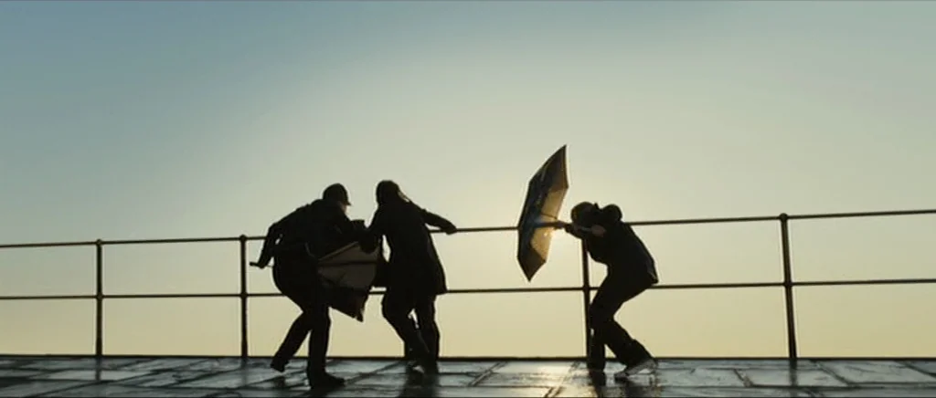 Sunshine movie still: wide — A metal railing runs horizontally across a damp pier surface against a clear sky. Three people struggle against strong…