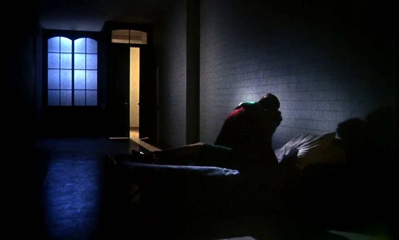 New York, New York (1977) movie still: wide — A stark cell features cinder block walls and a metal cot under a window. A man sits alone on the bed…