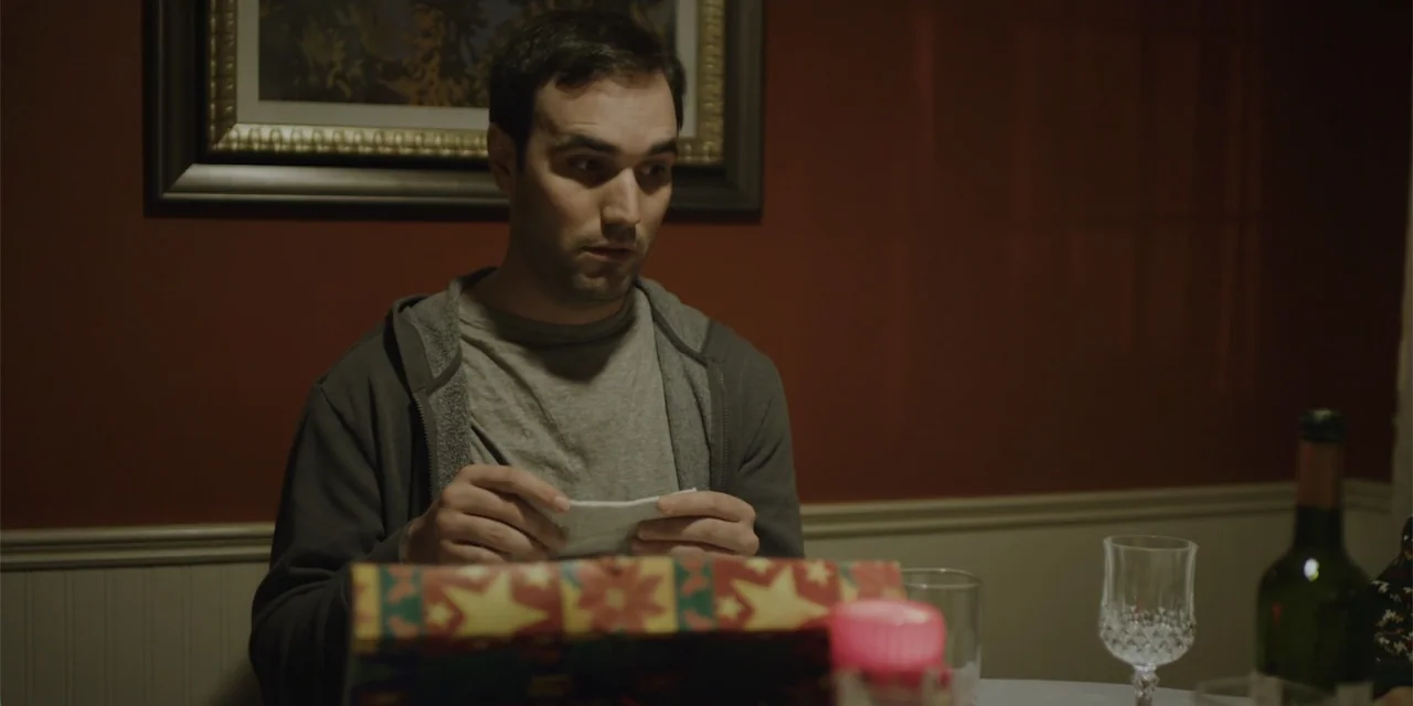 Lobos (2019) movie still: medium — A dimly lit room features a red wall with a framed print behind the man. A man in a hoodie sits at a table while…