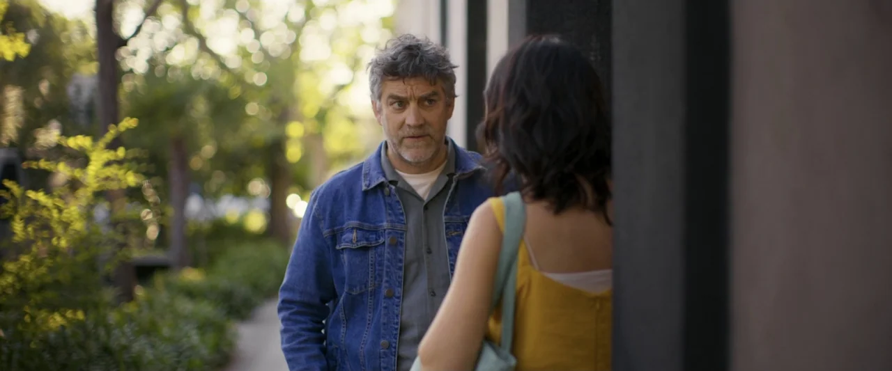 Dog Life (2024) movie still: medium — A sidewalk bordered by trees with a blurred background showing foliage and pavement. A man wearing a denim…
