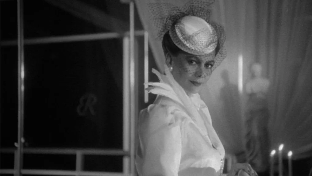 Under The Cherry Moon (1986) movie still: medium — An elegant woman stands in a dim room with sheer curtains and statue accents. She poses in white…