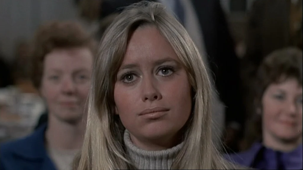 Straw Dogs (1971) movie still: closeup — A group of people sits in an indoor setting with muted tones. A young woman with long blonde hair looks…