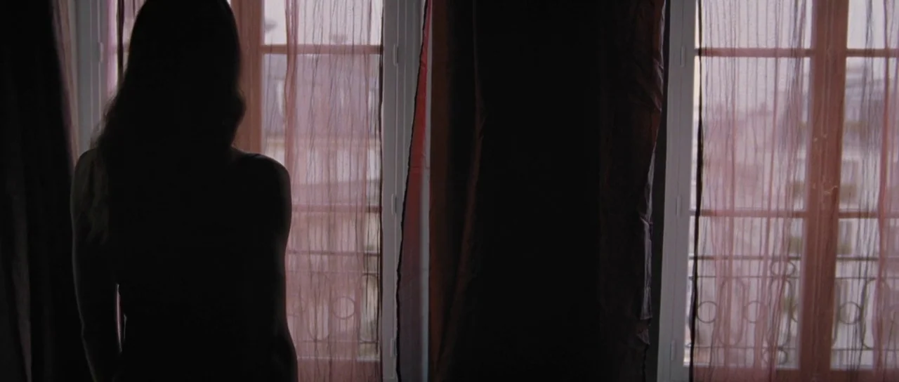 To The Wonder (2012) movie still: medium — A woman stands in front of a window covered with thin pink sheer curtains. The city exterior is partially…