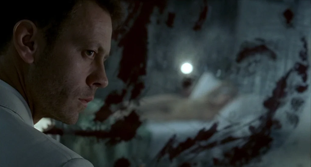 Anatomy of Hell (2004) movie still: closeup — A man in the foreground looks through a glass pane streaked with dark residue. Behind the glass, an…