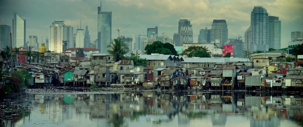 Samsara (2011) movie still: establishing — A dense shantytown of wooden and corrugated metal structures sits beside a polluted waterway. A modern…