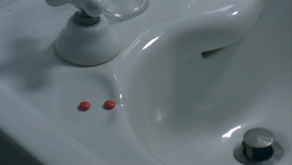 Sisters (1973) movie still: closeup — A white porcelain surface holds two small round red pills. The pills sit on the flat area between a chrome…