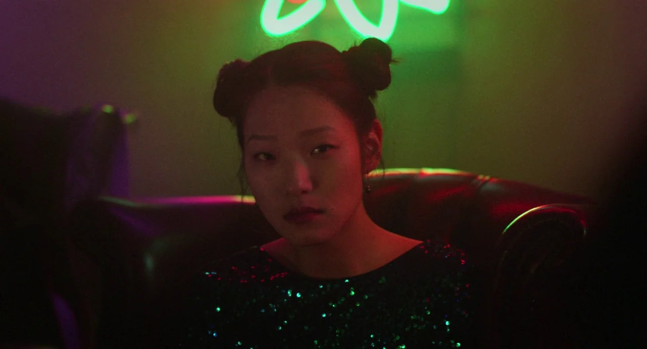 Return To Seoul (2022) movie still: closeup — A dark lounge interior features a woman sitting on a leather chair. A bright green neon geometric…