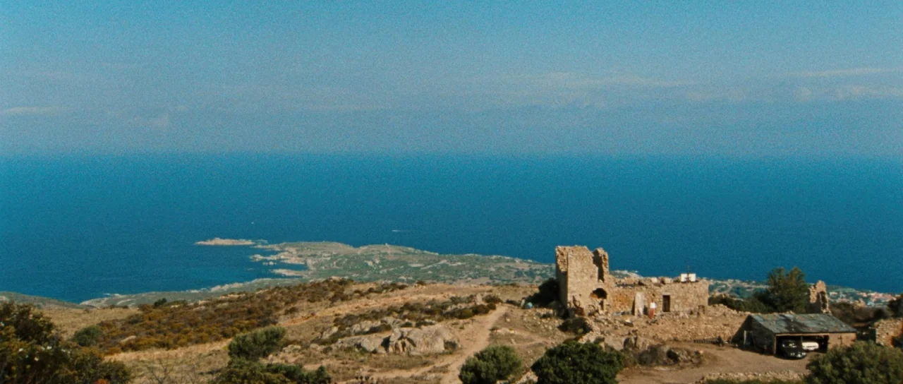 Let The Corpses Tan (2017) movie still: establishing — An ancient stone ruin sits on a dry, grassy hill overlooking a vast blue ocean. A small shed…