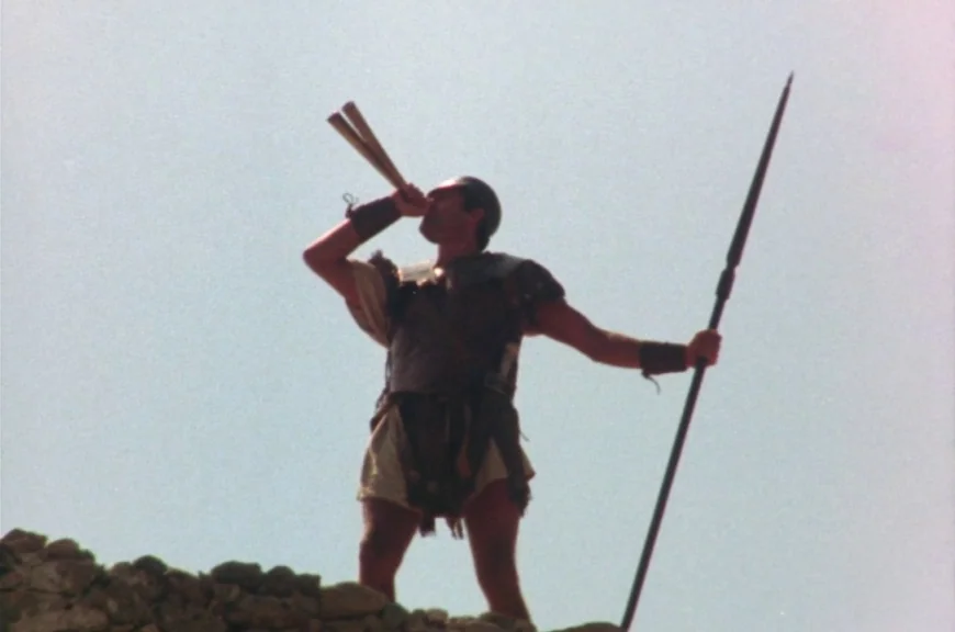 Sebastiane (1976) movie still: fullbody — A man stands atop a rocky ridge against a bright, hazy sky. He wears ancient leather armor and holds a…