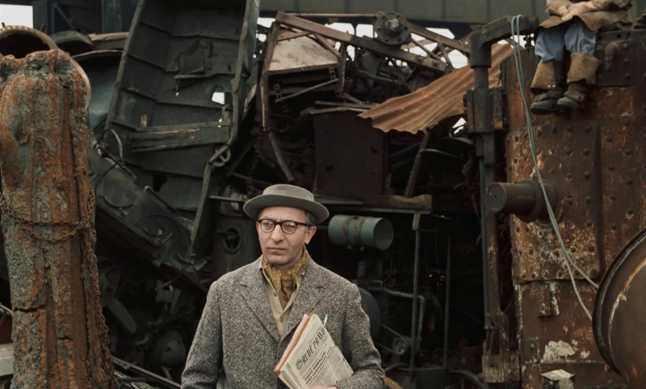 Larks On A String (1969) movie still: medium — A man stands before a massive, cluttered pile of rusted industrial machinery and metal. He holds a…