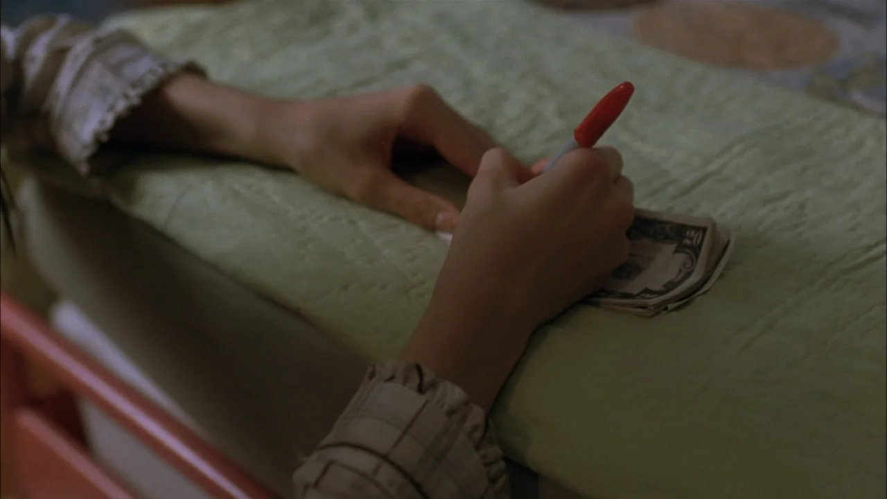 The Babysitters (2008) movie still: closeup — Hands rest on a pale green textile surface inside a room. The person holds a red marker to write on…