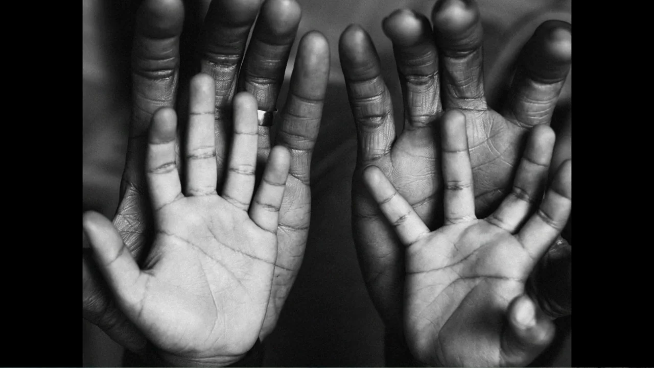 Between the World and Me (2020) movie still: closeup — A close-up view shows two large adult hands supporting two smaller child hands. The fingers…