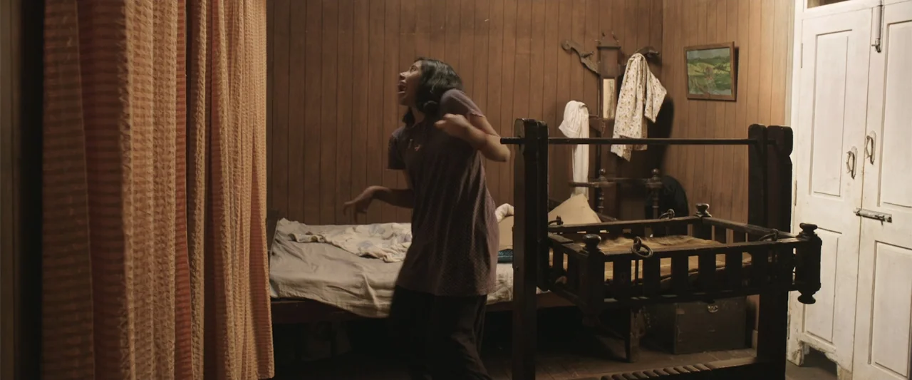 Peranbu (2019) movie still: fullbody — A wooden bedroom features a simple bed frame and a vintage wooden baby crib. A young woman stands in the…