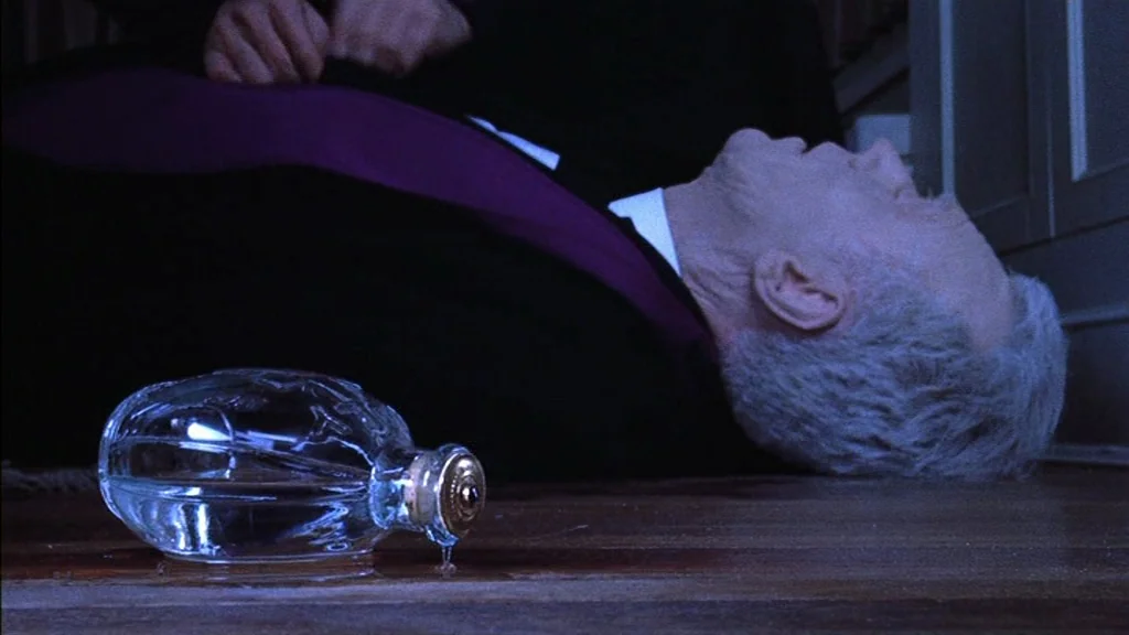 The Exorcist (1973) movie still: medium — A man lies prone on a wooden floor in a dimly lit room. A glass bottle with a gold stopper sits in the…